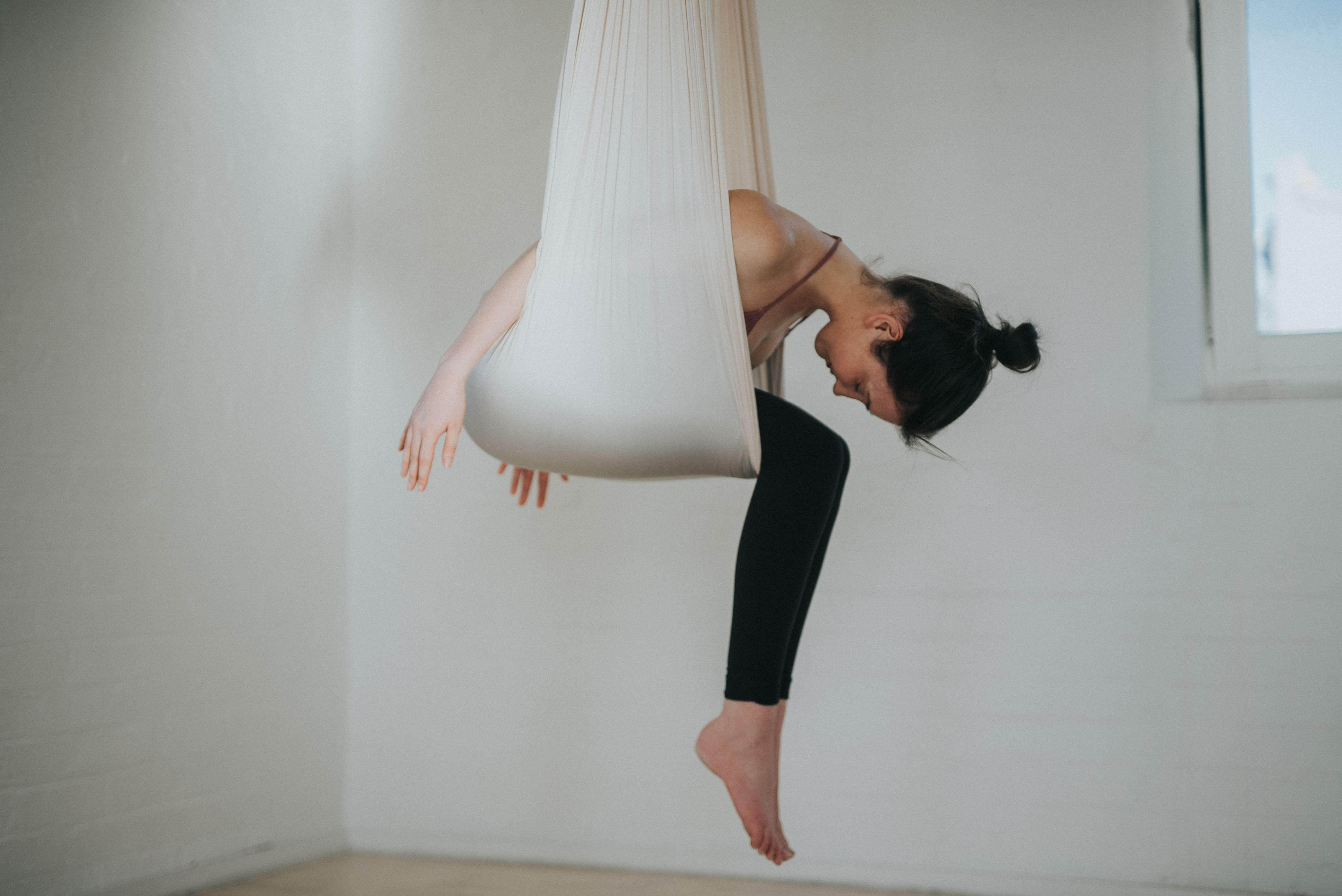 A woman resting in an aerial yoga hammock in a sunlit white studio