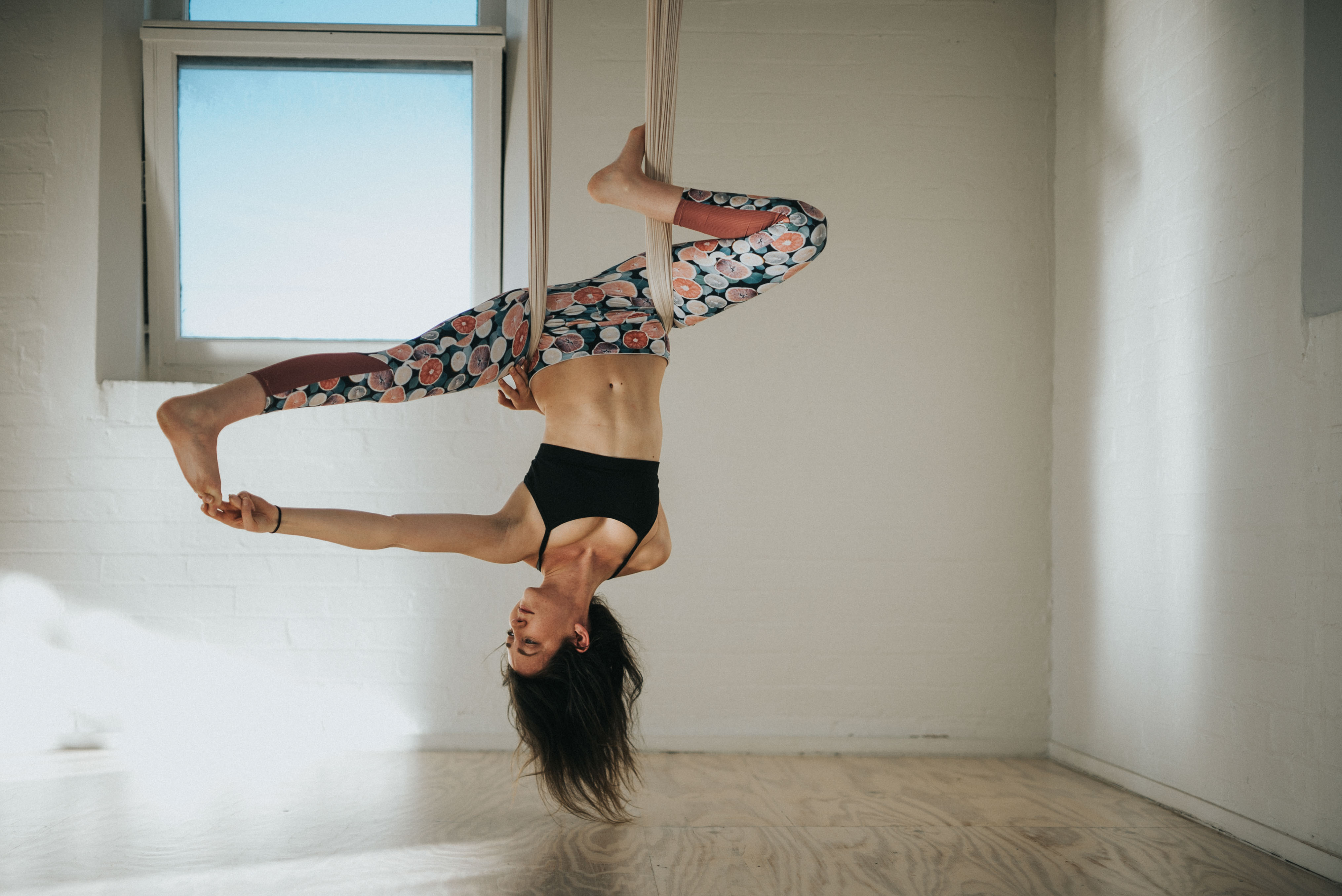 An aerial yogi inverted in a fabric hammock in a sunlit studio