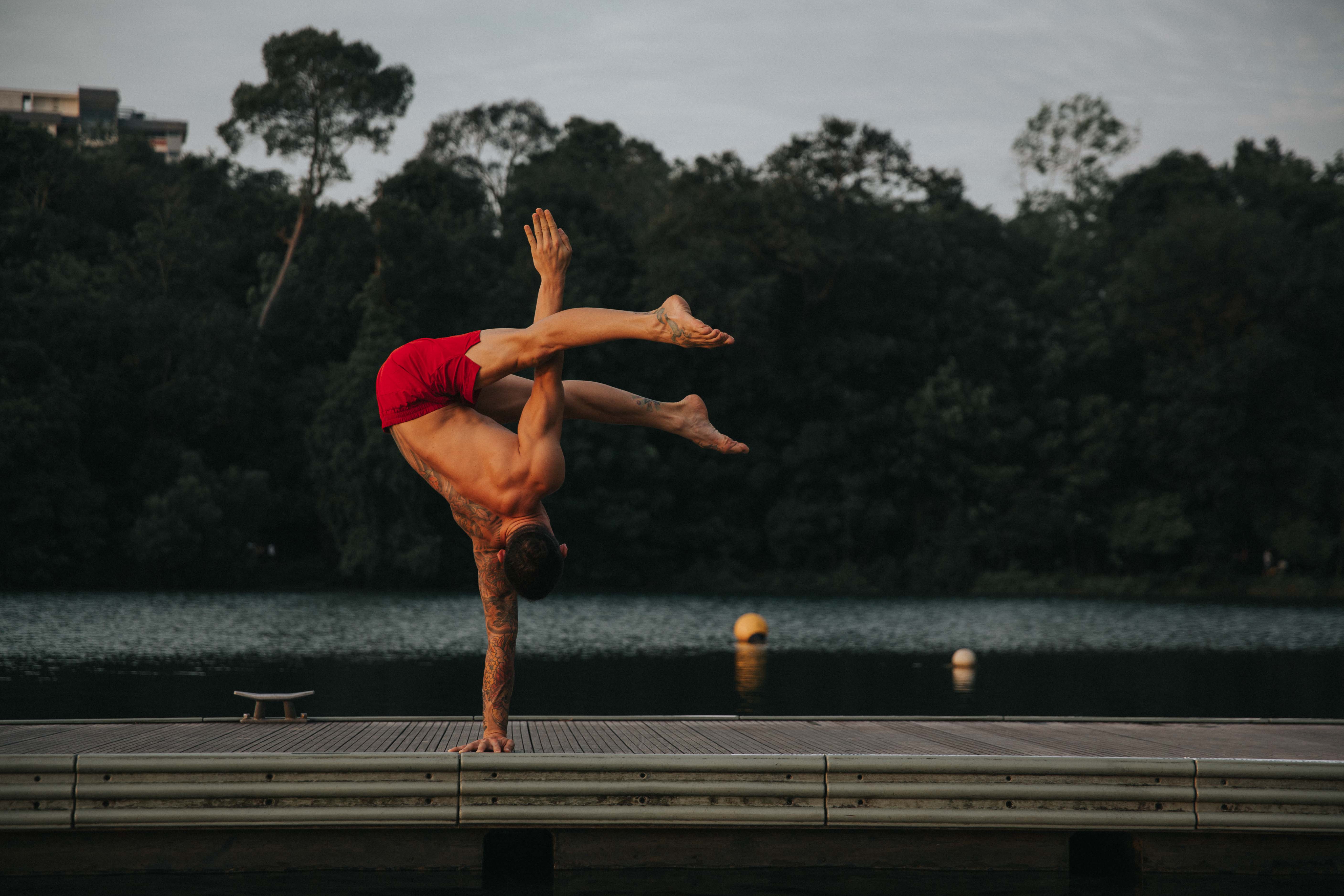 An athlete holding a one-arm handstand on a lakeside jetty at dusk