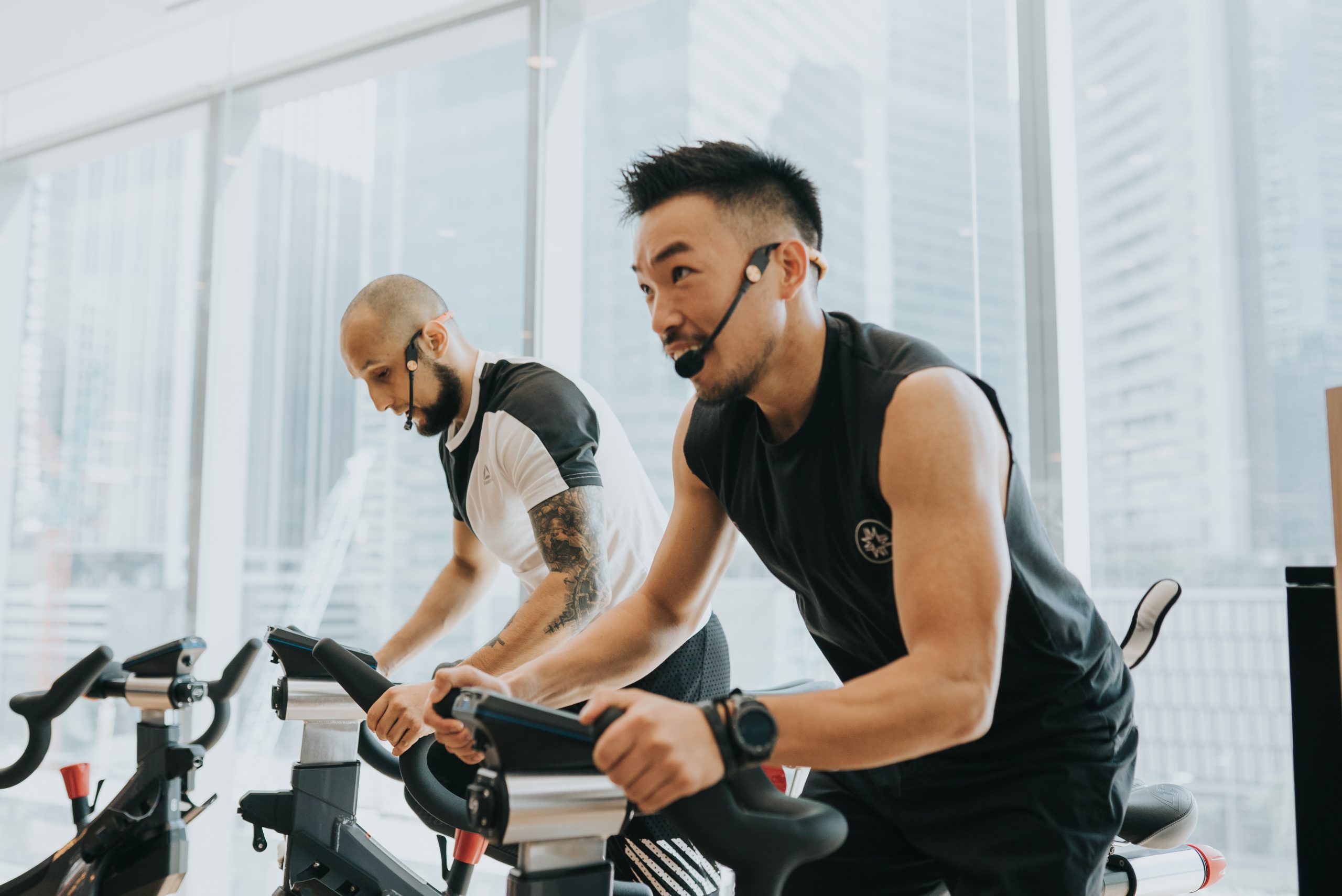 Two instructors leading an indoor cycling class by a floor-to-ceiling city window