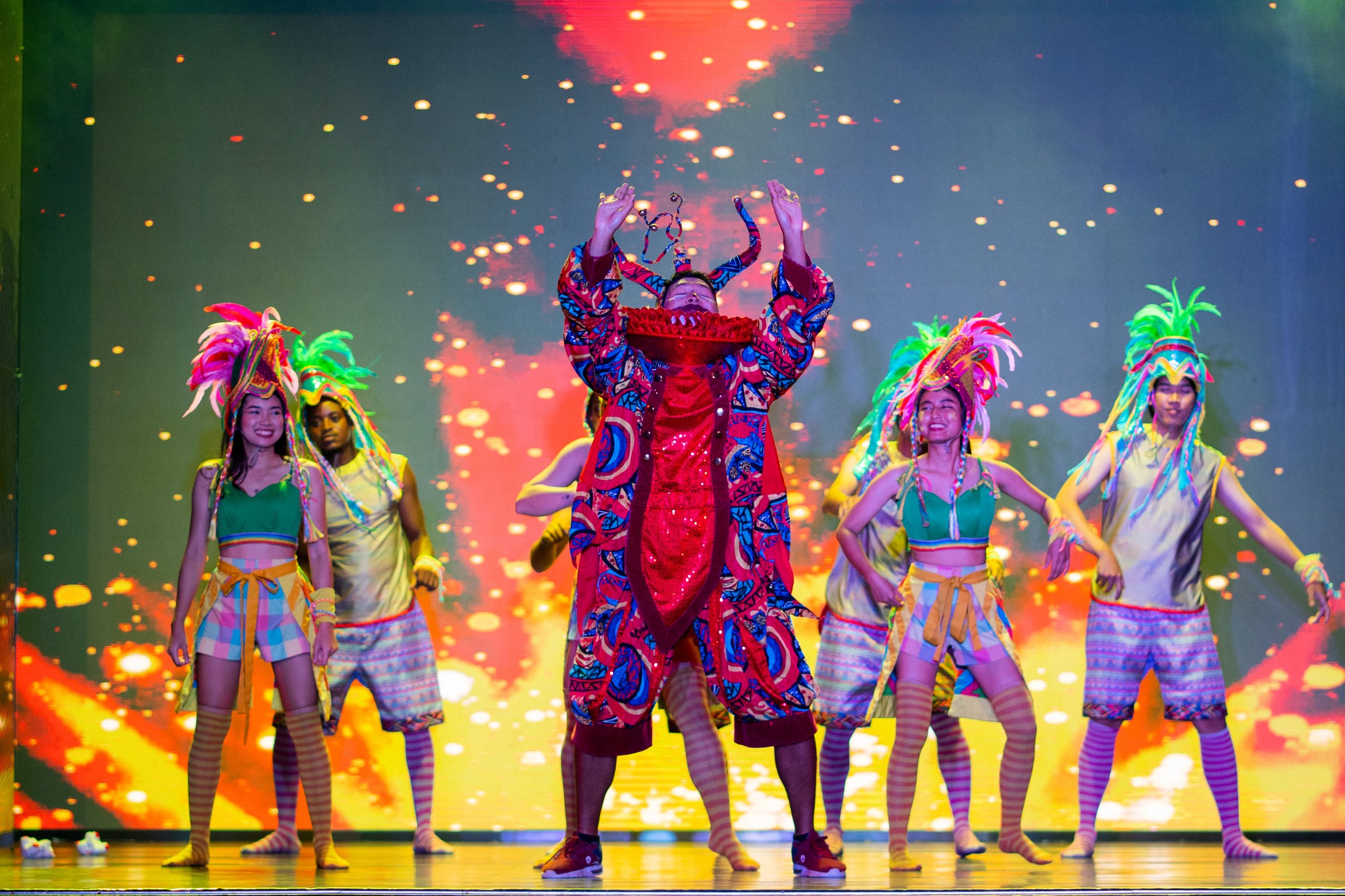 Performers in vibrant carnival costumes on a stage lit with fiery projections